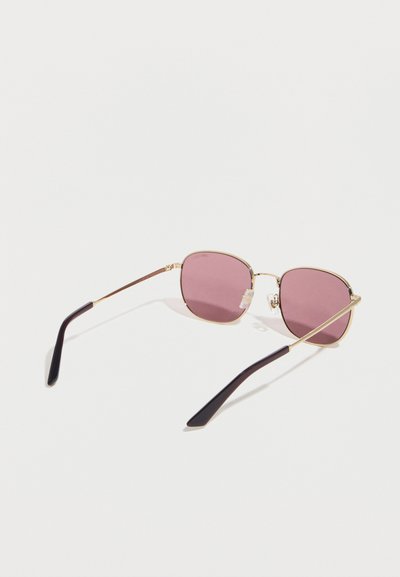 Γυαλιά ηλίου με χρυσή μεταλλική κορνίζα, τετράγωνα μοβ φακούς και μαύρες άκρες στα πλαινά, τοποθετημένα πάνω σε λευκή επιφάνεια.