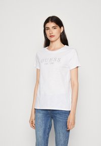 Balta medvilnės T-marškiniai su trumpomis rankovėmis, kuriuose viduryje yra metalinis "GUESS EST. 1981" logotipas. Modelis pozuoja vilkėdamas atsitiktinius mėlynus džinsus.