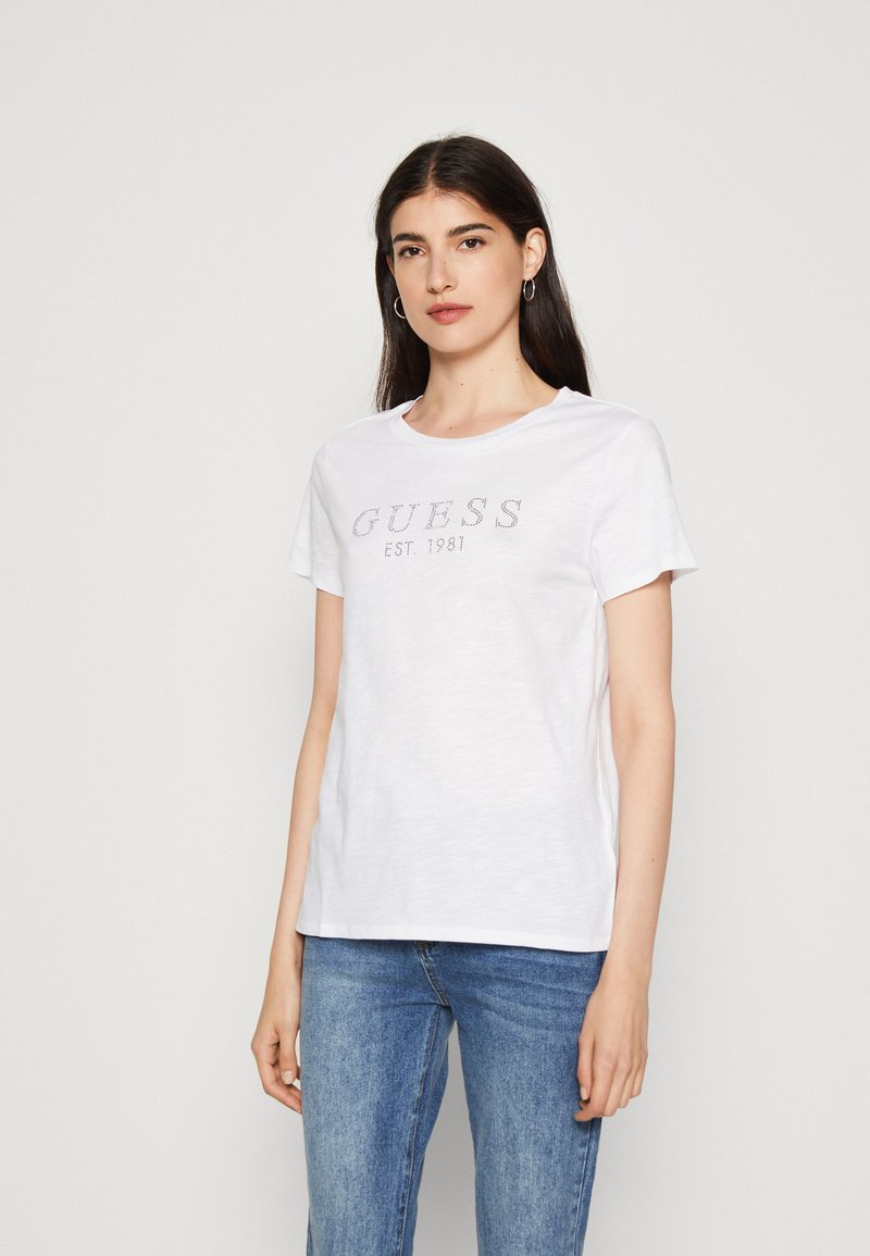 Balta medvilnės T-marškiniai su trumpomis rankovėmis, kuriuose viduryje yra metalinis "GUESS EST. 1981" logotipas. Modelis pozuoja vilkėdamas atsitiktinius mėlynus džinsus.