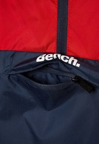 Veste coupe-vent rouge et marine avec une poche zippée à l'avant, logo « Bench. » et tissu lisse et léger avec des coutures contrastées.