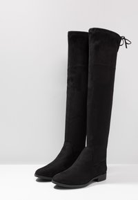 Black over-the-knee boots made from soft suede, featuring a rounded toe, flat sole, and an adjustable tie at the top for fit.
