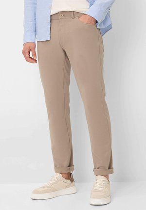 Beige Stretchpants mit tailliertem Schnitt, ausgestattet mit Seitentaschen und einem knöpfbaren Verschluss, kombiniert mit hellen Sneakers.
