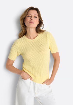 Frau mit hellbraunen Haaren trägt einen kurzärmligen gelben Strickpullover und weiße Hosen, steht mit den Händen in den Taschen vor einem weißen Hintergrund.