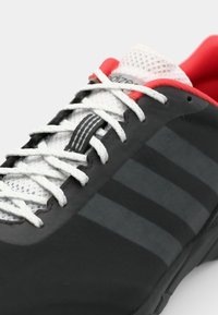 Black athletic shoe with white laces and mesh tongue; features red accents and grey stripes on the side; textured upper material.