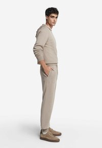 Beżowy sweter z kapturem i zwężane spodnie o miękkiej teksturze. Posiada boczne kieszenie i dopasowane mankiety. Połączone z jasnobrązowymi butami.