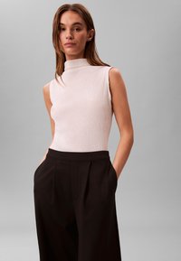 Frau mit glattem braunem Haar, die ein ärmelloses weißes Oberteil mit hohem Kragen und schwarze, weit geschnittene Hosen trägt, die Hände in den Taschen.