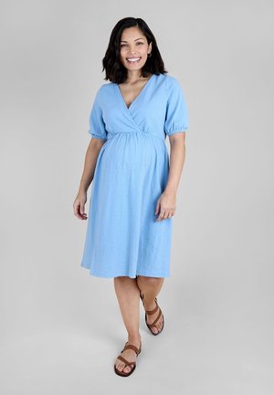 Uśmiechnięta ciężarna kobieta ubrana w jasnoniebieską sukienkę do kolan i brązowe sandały, stojąca na tle jednolitego szarego tła.