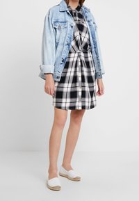Giacca di denim azzurro chiaro sopra un vestito a scacchi bianco e nero con bottoni. Espadrillas bianche completano l'outfit.