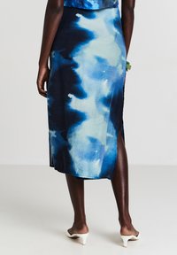 Rückansicht einer Person, die einen blau-weißen Batik-Midirock mit seitlichen Schlitzen und weißen High-Heel-Mules vor einem einfarbigen Hintergrund trägt.