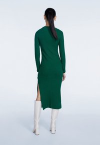 Robe à mailles côtelées vertes avec des manches longues, une fente sur le côté et une silhouette ajustée. Portée avec des bottes blanches montantes, exposée de dos.