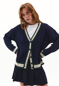 Cárdigan de punto en azul marino con acentos acanalados en verde y crema, cierre con botones en la parte delantera y ajuste holgado. Llevado sobre una camisa blanca.