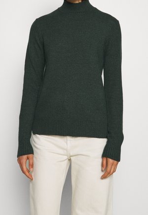 Person, die einen dunkelgrünen, gerippten Rollkragenpullover und cremeweiße Hosen trägt und vor einem einfachen, hellen Hintergrund steht.