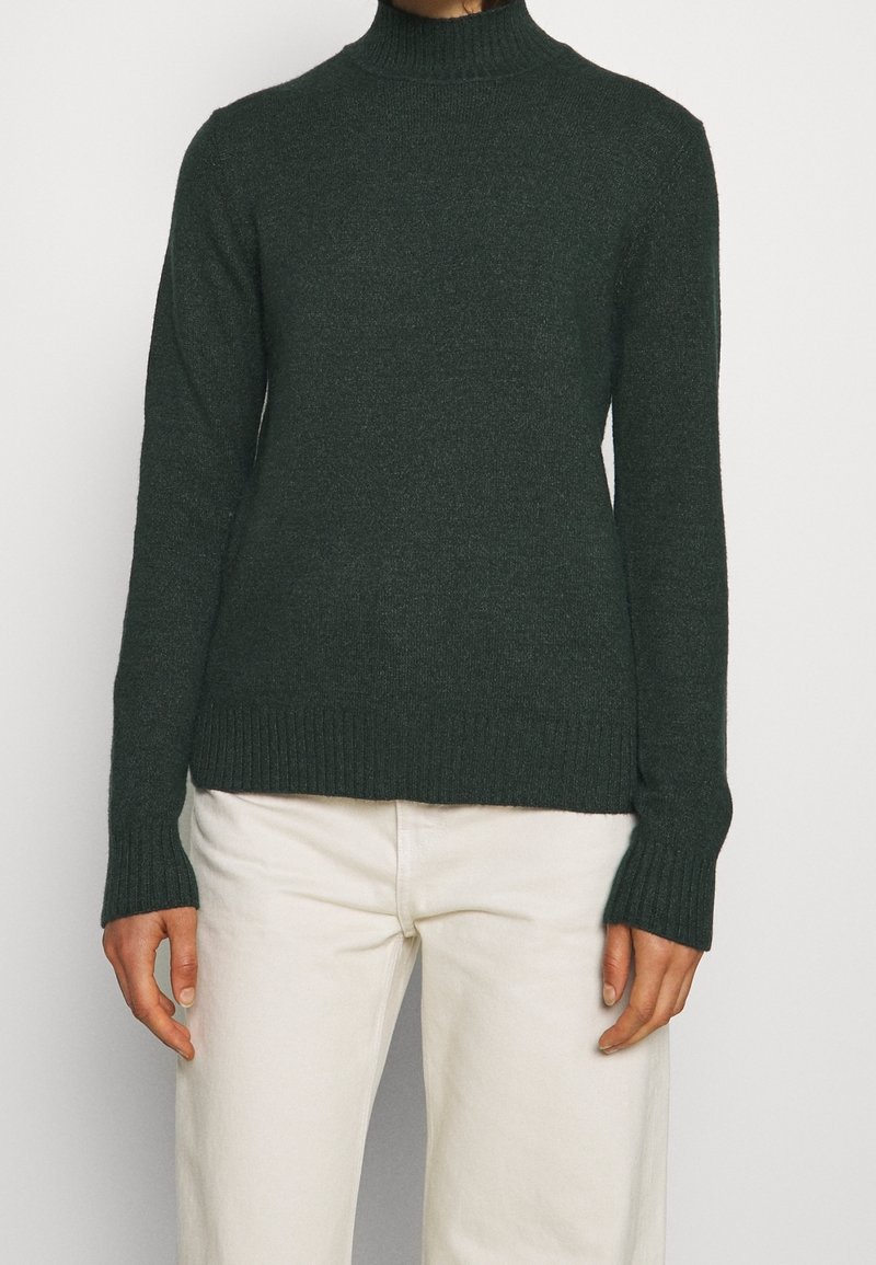 Personne portant un pull à col roulé côtelé vert foncé et un pantalon blanc cassé, debout devant un fond clair uni.