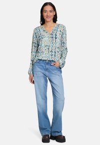 Femme souriante, portant un blouse à manches longues à motifs bleus, un jean bleu clair et des chaussures à plateforme sombres, se tenant avec une main dans la poche.