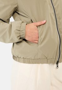 Letthvægts, beige bomberjakke med elastiske manchetter og kant, som har en frontlynlås og sidelommer. Glat, struktureret stof.
