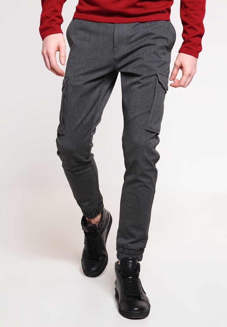 Homme portant un pantalon cargo jogging gris foncé avec poches latérales, des baskets noires, et un t-shirt à manches longues rouge, marchant vers l'avant sur un fond uni.