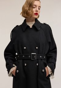 Czarny trencz z mieszanki wełny, z dwurzędowym zapięciem, srebrnymi guzikami, paskiem w talii oraz kieszeniami z patkami. Posiada kołnierz i mankiety z detalem.