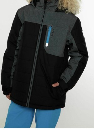 Person iført en sort og mørkegrå jakke med en blå lynlåslomme og lyseblå bukser, stående med den ene hånd i lommen.