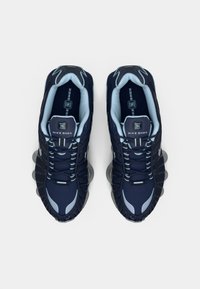 Pāris tumši zilu un gaiši zilu Nike Shox sporta apavu, skatoties no augšas uz vienkārša balta fona.