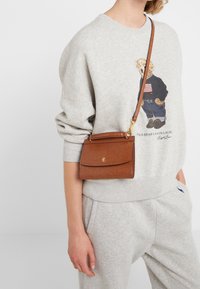 Hnědá kožená crossbody taška s texturovaným povrchem, klopovým uzávěrem a logem ve zlatém odstínu. Nastavitelný popruh umožňuje všestranné nošení.