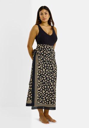 Femme portant un haut noir sans manches et une longue jupe noire avec un imprimé léopard beige et une bordure rayée, debout pieds nus sur fond blanc.