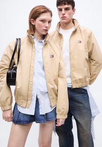 Δύο νεαροί ενήλικες φορούν μπεζ μπουφάν Tommy Hilfiger με φερμουάρ, η γυναίκα κρατά μια μαύρη τσάντα, στέκονται μπροστά σε ένα απλό λευκό φόντο.