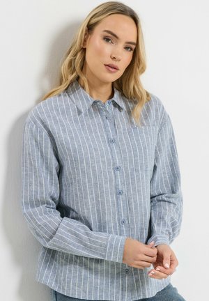 Lyshåret kvinde iført en lyseblå, langærmet skjorte med hvide lodrette striber, stående mod en hvid baggrund.