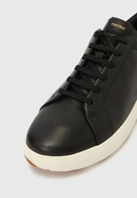 Μαύρο δερμάτινο αθλητικό παπούτσι με επίπεδοι κορδόνια, στρογγυλερή μύτη, λευκή λαστιχένια σόλα και  διακριτικό λογότυπο στη φτέρνα. Λεπτή υφή και κομψός σχεδιασμός.