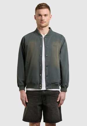 Mann, der eine dunkelgrüne, geknöpfte Bomberjacke über einem weißen Hemd und schwarzen Jeansshorts trägt und vor einem schlichten grauen Hintergrund steht.