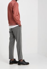 Man wearing gray trousers, black dress shoes, white shirt, and pinkish jacket, standing with hands in pockets against plain background.