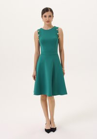 Teal obleka fit-and-flare z okroglim izrezom, zlatimi gumbi na ramenih in gladkim materialom. Črne špicaste petke dopolnjujejo videz.