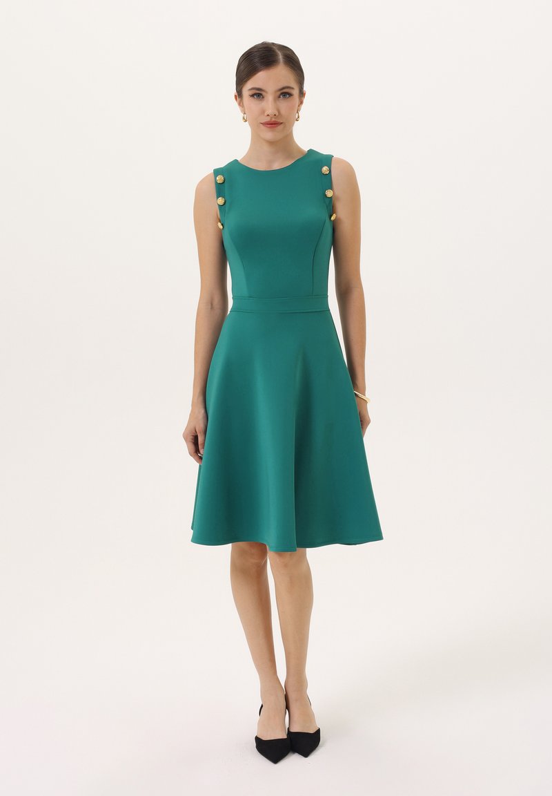 Teal obleka fit-and-flare z okroglim izrezom, zlatimi gumbi na ramenih in gladkim materialom. Črne špicaste petke dopolnjujejo videz.