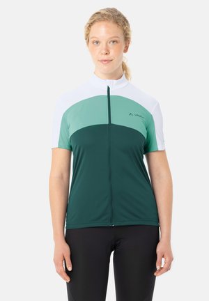 Vrouw staat rechtop en kijkt naar voren, draagt een korte-mouwen wielrennersshirt met groene en witte kleurvlakken en zwarte wielershorts.