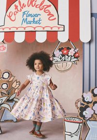 Noor tüdruk värvikirevas kleidis seisab punase-valge triibulise varikatuse all, ümberringi on multikatega lilleturulised dekoratsioonid.