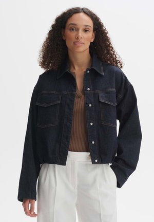 Dark denim jacket featuring two front pockets, button closure, and a relaxed fit. Worn over a brown ribbed top, paired with white trousers.