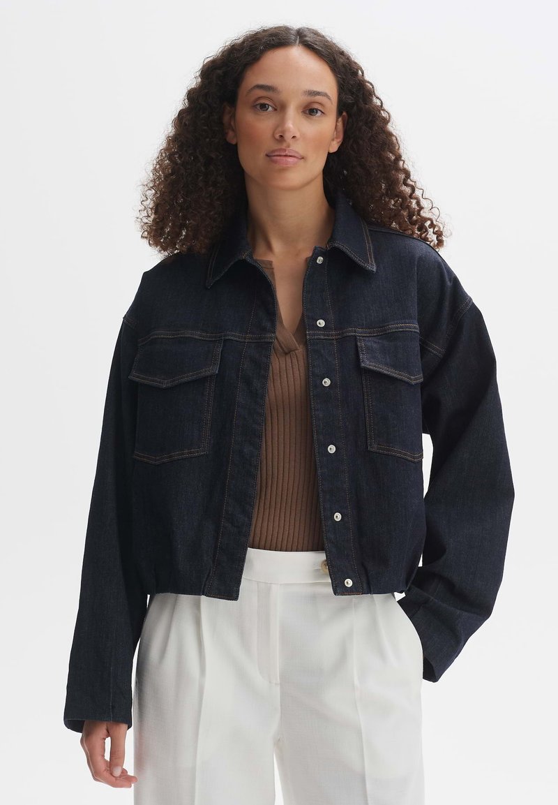 Veste en denim foncé dotée de deux poches avant, d'une fermeture à boutons et d'une coupe décontractée. Portée par-dessus un top marron à côtes, associée à un pantalon blanc.