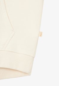 Krämmfärgad sweatshirtärm med ribbad manschett och sidoficka. Har en liten, beigefärgad tygskylt sydd vid kanten.