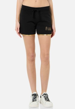 Weibliches Model trägt schwarze Shorts mit goldfarbenem Saum und „Boxeur Des Rues“-Logo, kombiniert mit weißen Sneakers mit schwarzen Schnürsenkeln.