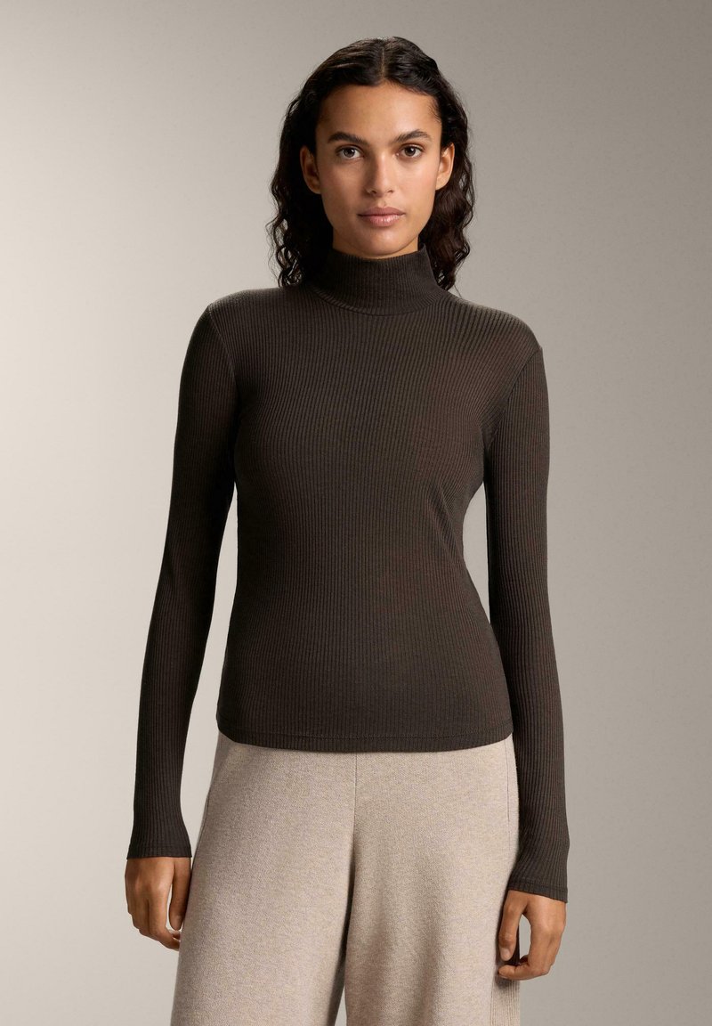 Rippstrick-Turtleneck-Oberteil in Dunkelbraun, figurbetonter Schnitt mit langen Ärmeln, weicher Textur. Kombiniert mit hellbeigen Hosen, minimalistischem Stil.