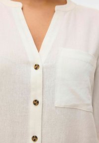 Camicia bianca di lino con scollatura a V, dotata di taschino sul petto e bottoni marroni lungo la parte frontale. Tessuto leggero e materiale traspirante.