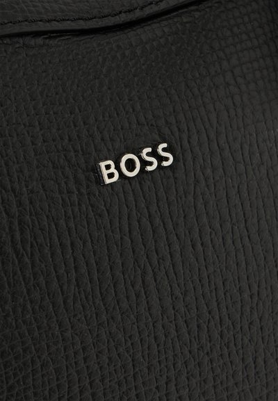 Gros plan sur un sac en cuir noir grainé orné d’un petit logo BOSS argenté.