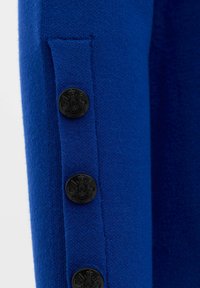 Blauwe wollen stof heeft een rechtse mouw met een verticale zak en vier ronde zwarte knoppen met een wapen.
