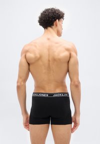 Άντρας με σγουρά μαλλιά, μυώδης πλάτη, φοράει μαύρα στενά εσώρουχα με λάστιχο μέσης "JACK & JONES", στέκεται μπροστά σε ανοιχτόχρωμο απλό φόντο.