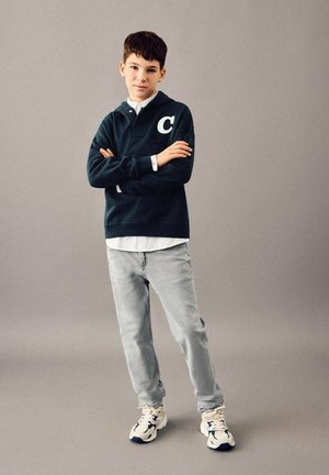Poika seisoo käsivarret ristissä päällään tumma huppari, jossa on kirjain "C", valkoinen paita, vaaleat farkut ja valkoiset tennarit harmaata taustaa vasten.