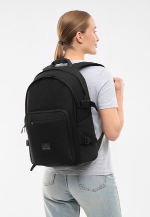 Black backpack with padded straps, front zip pocket, and logo patch; features smooth texture and streamlined design. Worn on one shoulder.