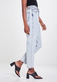 Jeans taille haute bleu clair avec une coupe décontractée, présentant une texture dégradée, des poches avant et arrière, et une étiquette en cuir beige à la taille. Talons aiguilles noirs à lanières.
