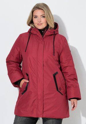 Donna che indossa un cappotto invernale rosso con cappuccio e tasche bordate di nero, in piedi davanti a uno sfondo chiaro e uniforme, mano in tasca.