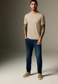 Kreminė trumpomis rankovėmis marškinėliai, mėlynos džinsinės kelnės ir kreminės zomšinės sportbačiai. Modelis demonstruoja tatuiruotes ant rankų. Neutralus fonas pabrėžia drabužių spalvas.