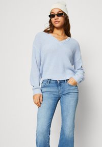 Lyseblå, strikket genser med bølgete V-hals og lange ermer, parret med blå denim jeans. Modellen har på seg briller og en hvit lue.