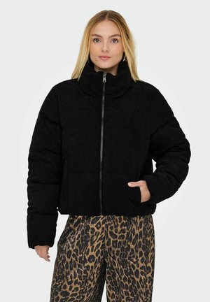 Femme aux cheveux blonds portant une doudoune noire zippée et un pantalon à imprimé léopard, une main dans la poche de la veste.